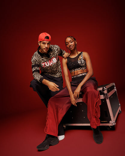 A male and a female model pose together in a studio setting with a solid dark red background and sitting on an equipment case. The man wears a red backwards hat, a leopard-print long-sleeve shirt with 'ZUMBA' printed on the front, black pants, and white sneakers. The female wears a leopard print bra with asymmetrical mesh overlay and black and red gradient pants with black shoes.