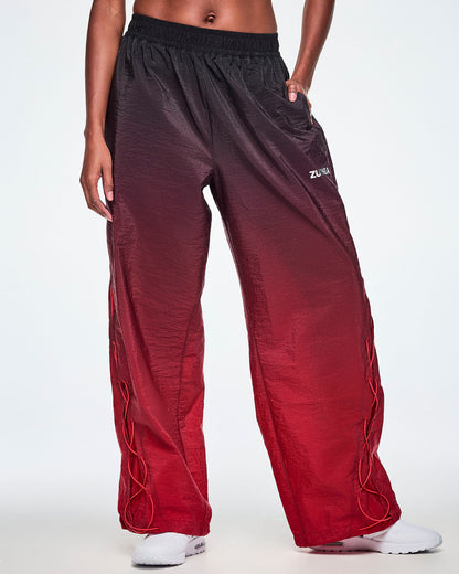 Front view of a female model wearing loose, wide-leg gradient pants that fade from black at the top to deep red at the bottom and showing the lower leg panel with adjustable criss-cross lace up detail and paired with white shoes.