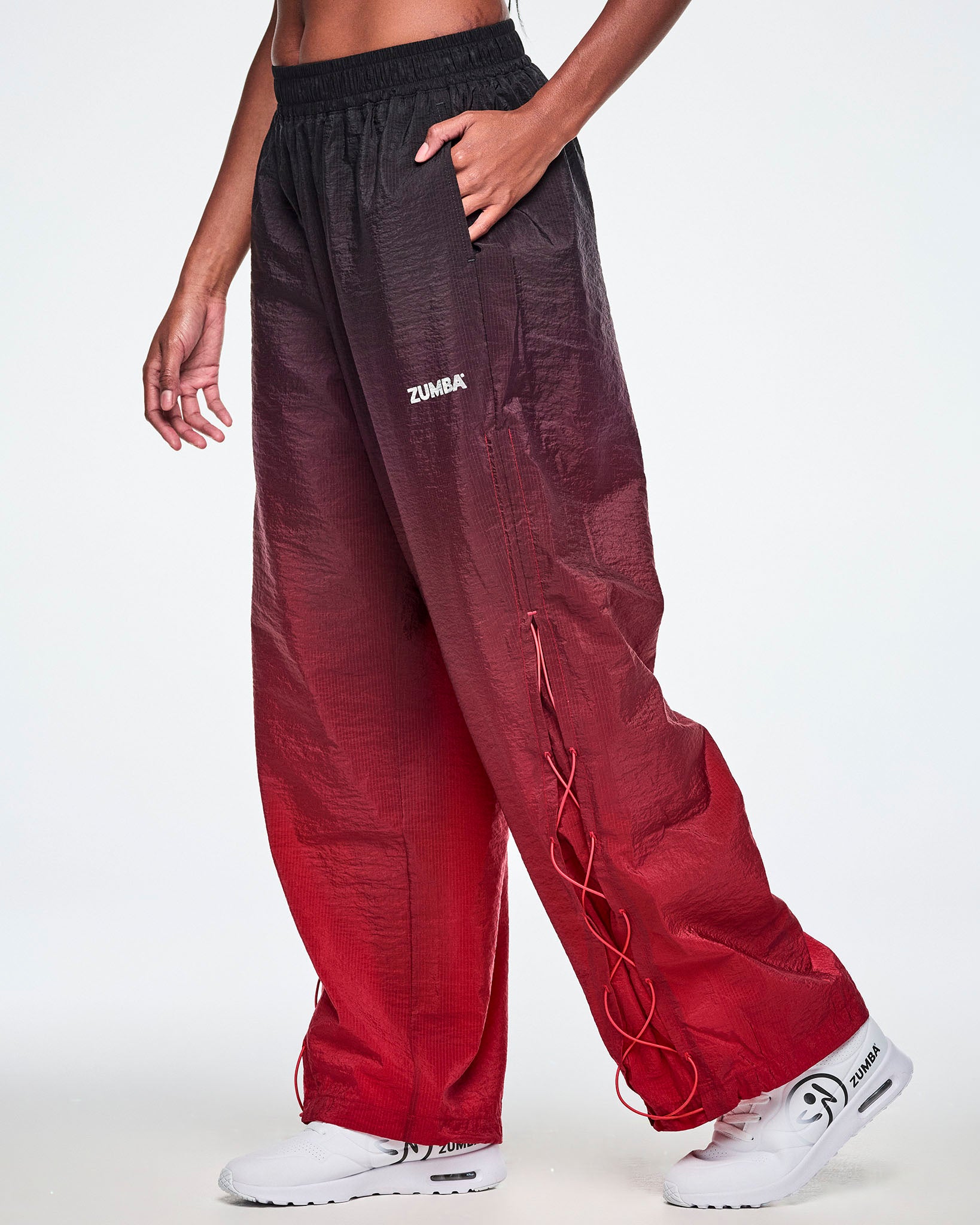 A female model wearing loose, wide-leg gradient pants that fade from black at the top to deep red at the bottom and showing the lower leg panel with adjustable criss-cross lace up detail and paired with white shoes.