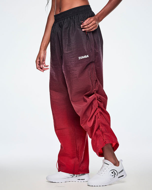 Side view of a female model wearing red gradient pants and pulling on the bungee cord toggle in pocket to adjust length of pants and paired with white shoes.