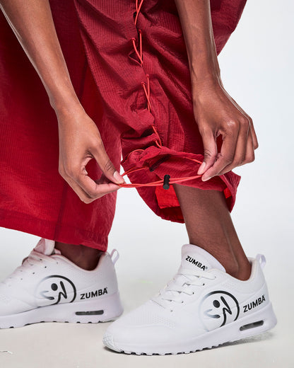 A close-up view of a female model wearing black and red gradient pants and pulling on the adjustable criss-cross lace up detail from the ankles and paired with white Zumba shoes.