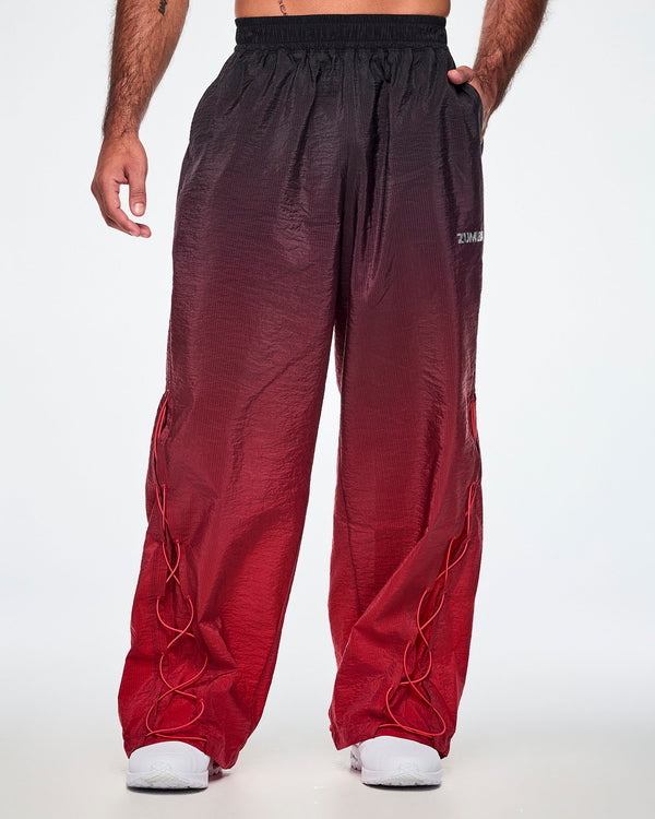 Front view of a male model wearing loose, wide-leg gradient pants that fade from black at the top to deep red at the bottom and showing the lower leg panel with adjustable criss-cross lace up detail and paired with white shoes.