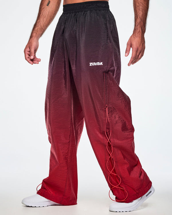 A male model wearing loose, wide-leg gradient pants that fade from black at the top to deep red at the bottom and showing the lower leg panel with adjustable criss-cross lace up detail and paired with white shoes.