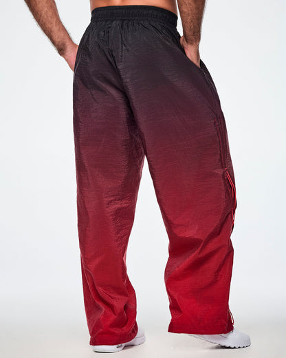 Back view of a male model wearing loose, wide-leg black to red gradient pants, paired with white shoes.