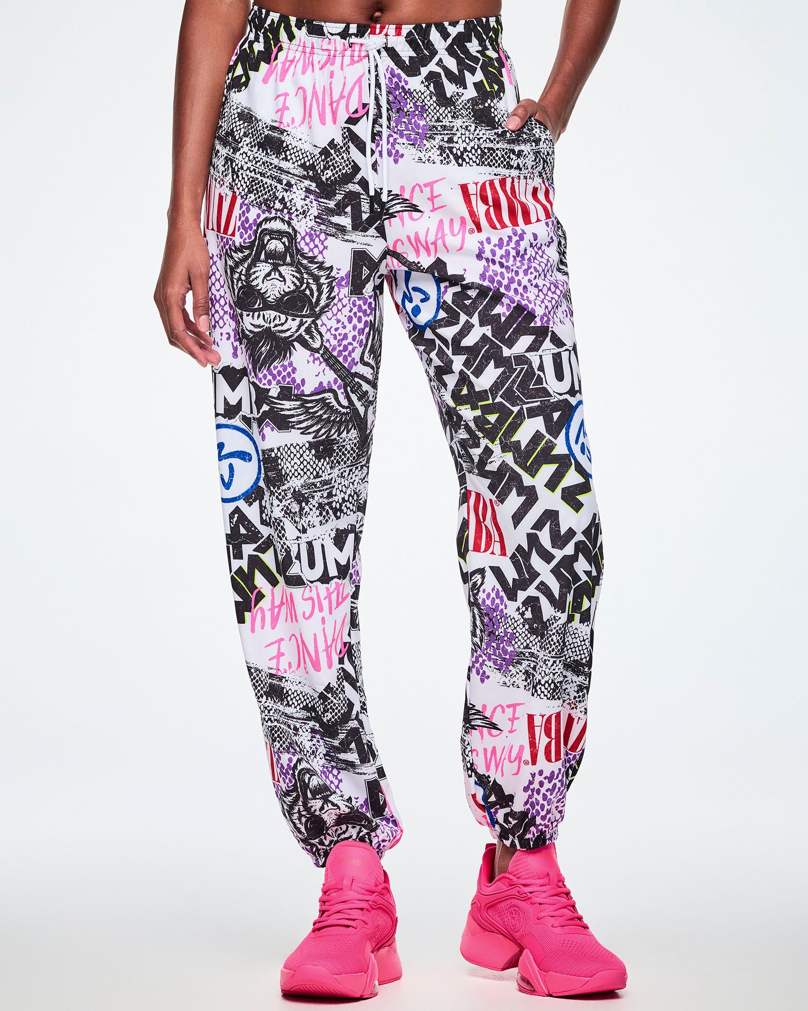 Front view of a female model, with a hand in her pocket of the white woven pants with an all-over ripped newspaper like pattern with black, white and red ZUMBA text and pink Dance this way text along with other Zumba rock graphics, Zumbitos and purple and black snakeskin patterns, paired with pink shoes.