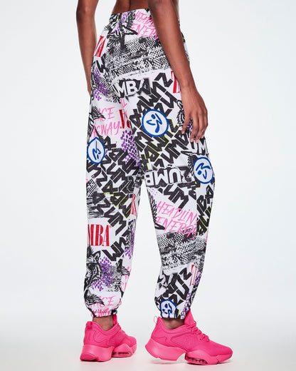 Back view of a female model wearing white woven pants with an all-over ripped newspaper like pattern with black, white and red ZUMBA text and pink Dance this way text along with other Zumba rock graphics, Zumbitos and purple and black snakeskin patterns, paired with pink shoes.