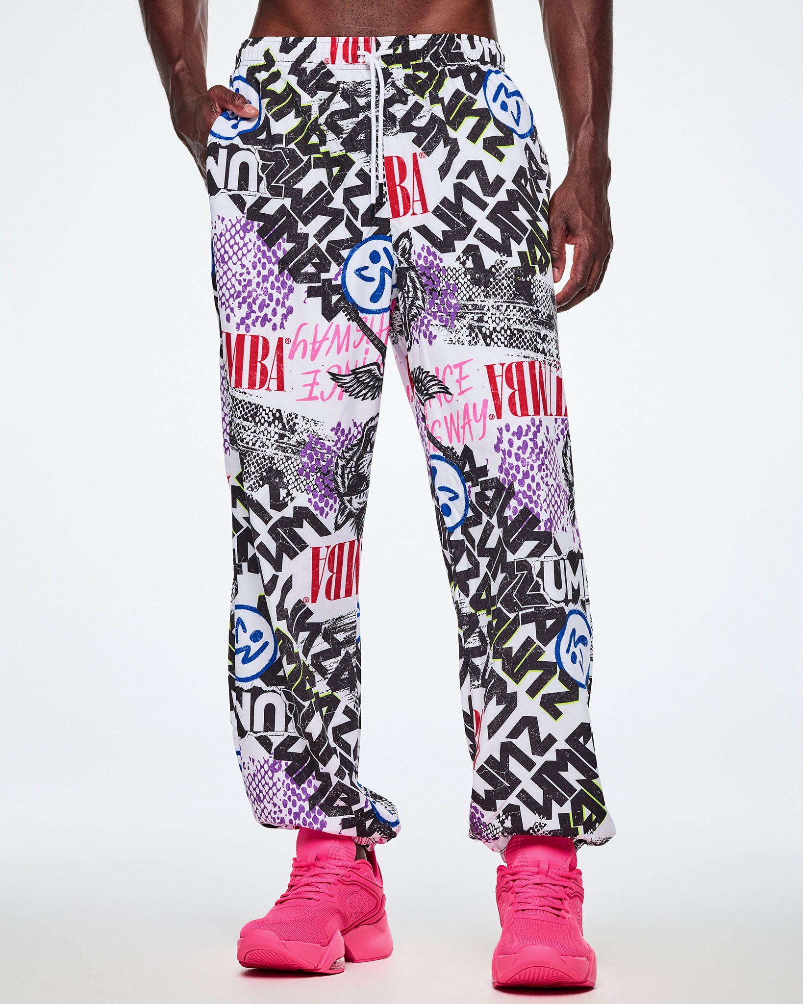 A male model with one hand in his pocket of the white woven pants with an all-over ripped newspaper like pattern with black, white and red ZUMBA text and pink Dance this way text along with other Zumba rock graphics, Zumbitos and purple and black snakeskin patterns, paired with pink shoes.