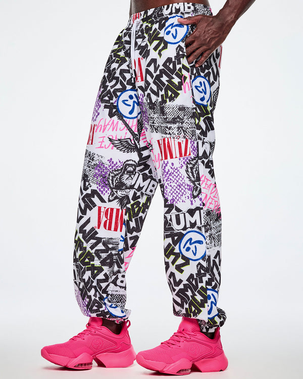 A male model wearing white woven pants with an all-over ripped newspaper like pattern with black, white and red ZUMBA text and pink Dance this way text along with other Zumba rock graphics, Zumbitos and purple and black snakeskin patterns, paired with pink shoes.