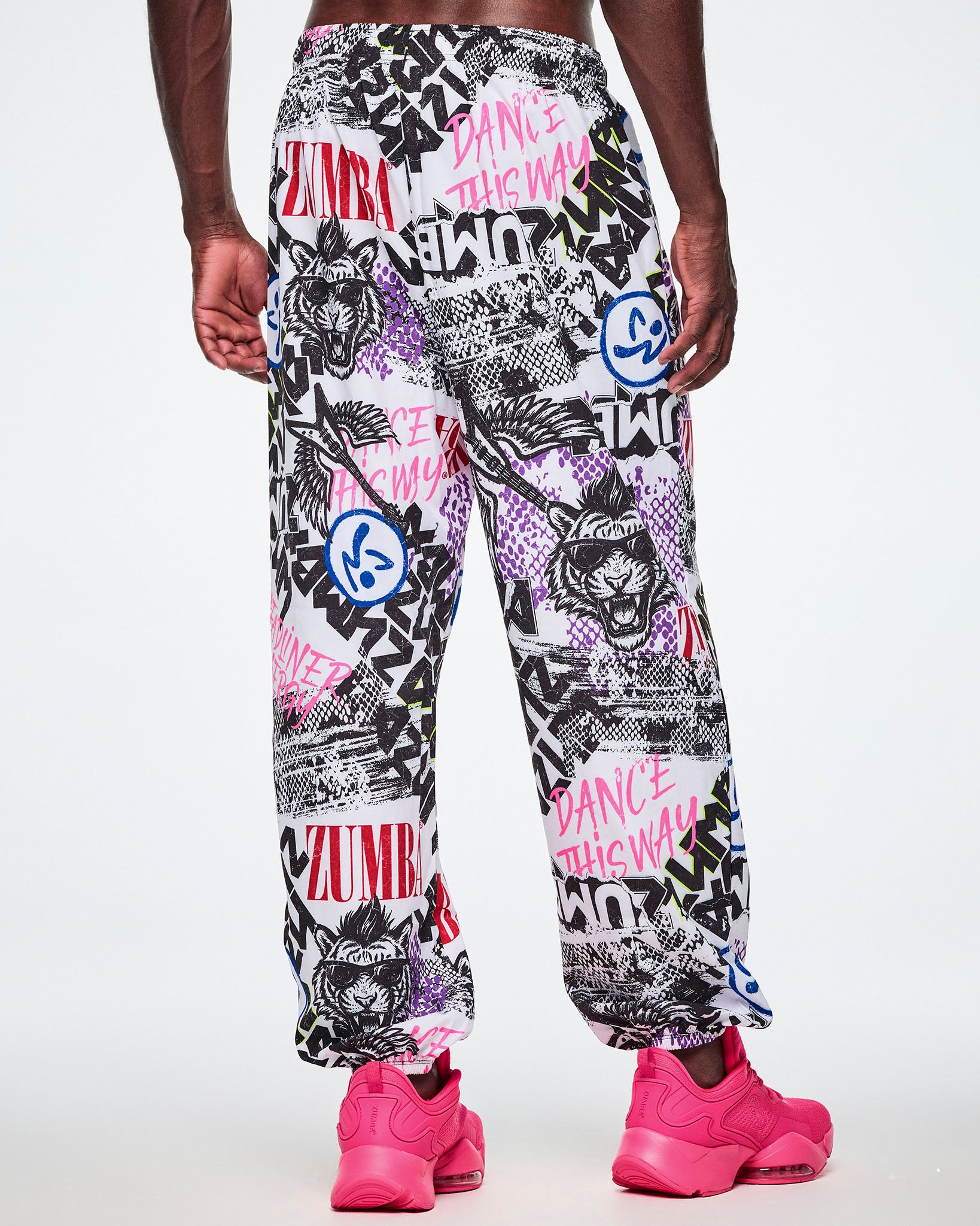 Back view of a male model wearing white woven pants with an all-over ripped newspaper like pattern with black, white and red ZUMBA text and pink Dance this way text along with other Zumba rock graphics, Zumbitos and purple and black snakeskin patterns, paired with pink shoes.