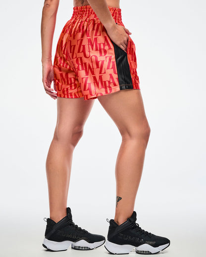 Back view of a female model wearing orange boxing shorts with an allover red “ZUMBA” monogram, one hand in the pocket, and a black side stripe.