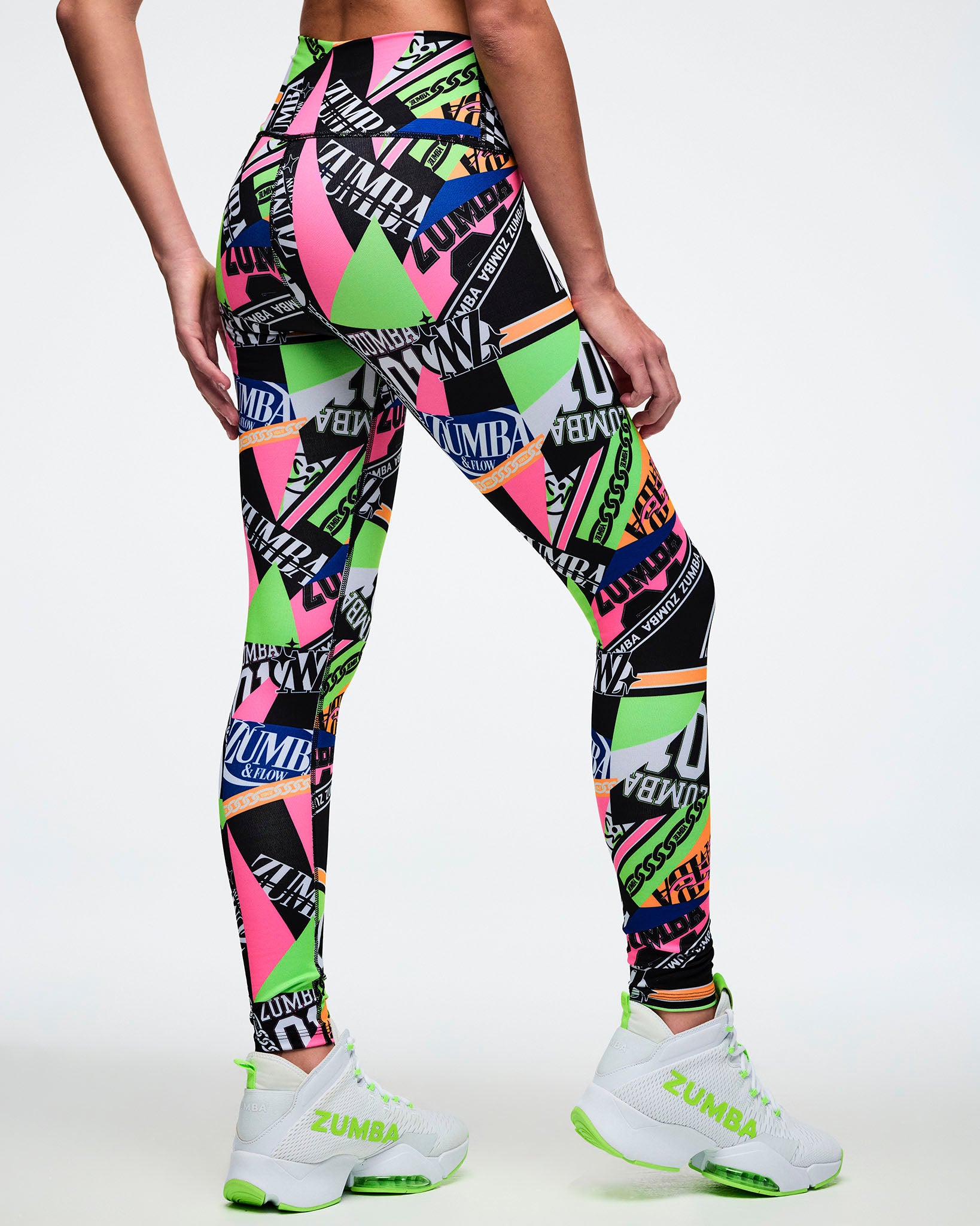 Back view of a female model wearing multi-colored Zumba leggings with a geometric pattern paired with matching green and white shoes.