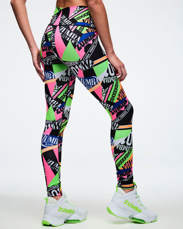 Back view of a female model wearing multi-colored Zumba leggings with a geometric pattern paired with matching green and white shoes.