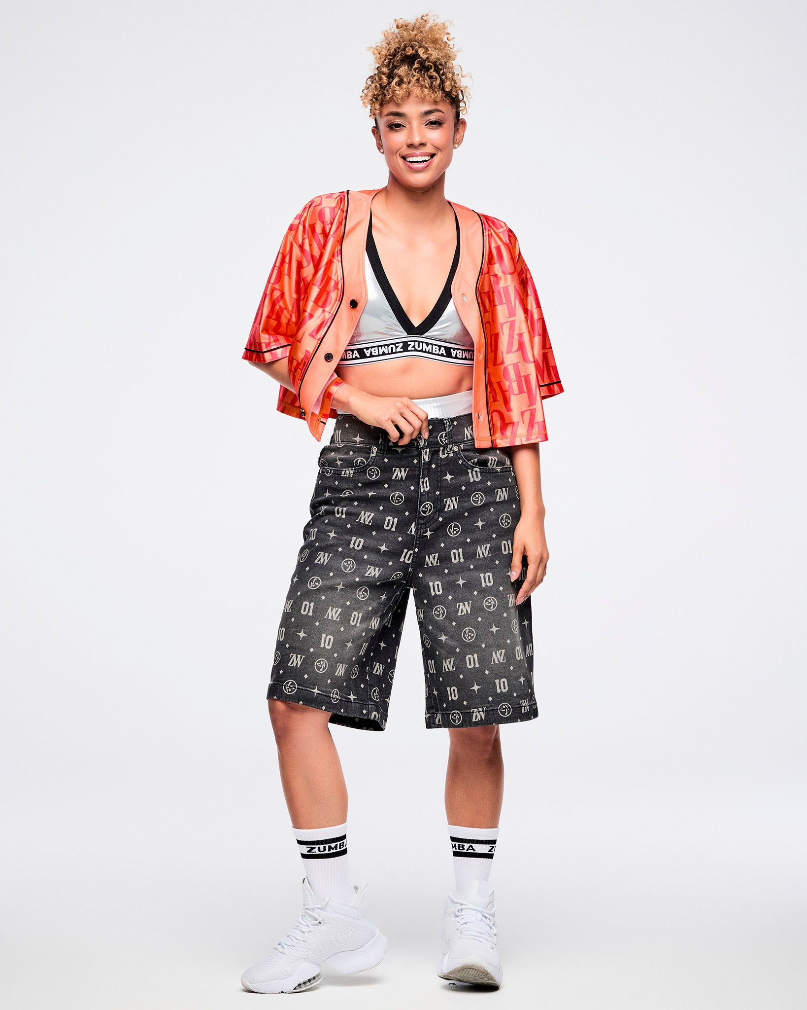 Female model wearing an open red button-up top over a silver bra, pulling up black denim jorts with one hand that features ZW, “01,” stars, dots, and Zumbito motifs, paired with black-and-white socks and shoes.