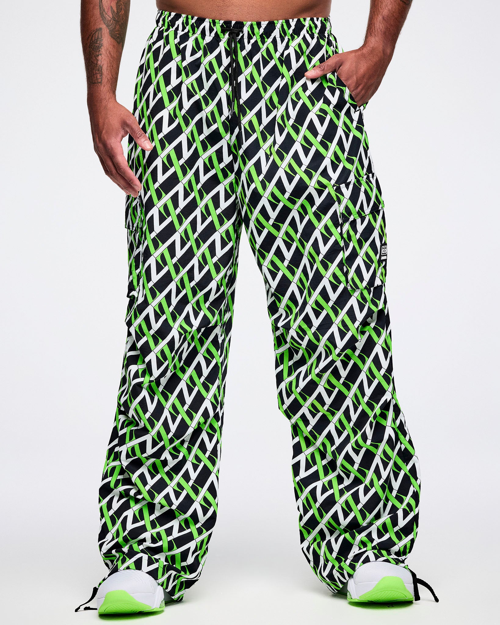 Front view of a male model wearing black cargo pants with an allover black, white and green “Z” crisscross pattern, left hand in his pocket, paired with black-and-white shoes.