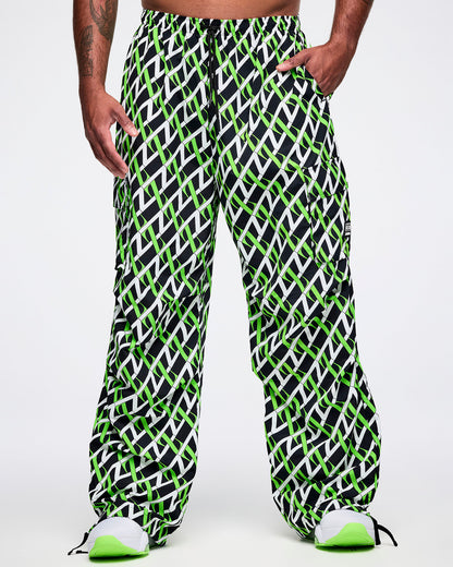 Front view of a male model wearing black cargo pants with an allover black, white and green “Z” crisscross pattern, left hand in his pocket, paired with black-and-white shoes.
