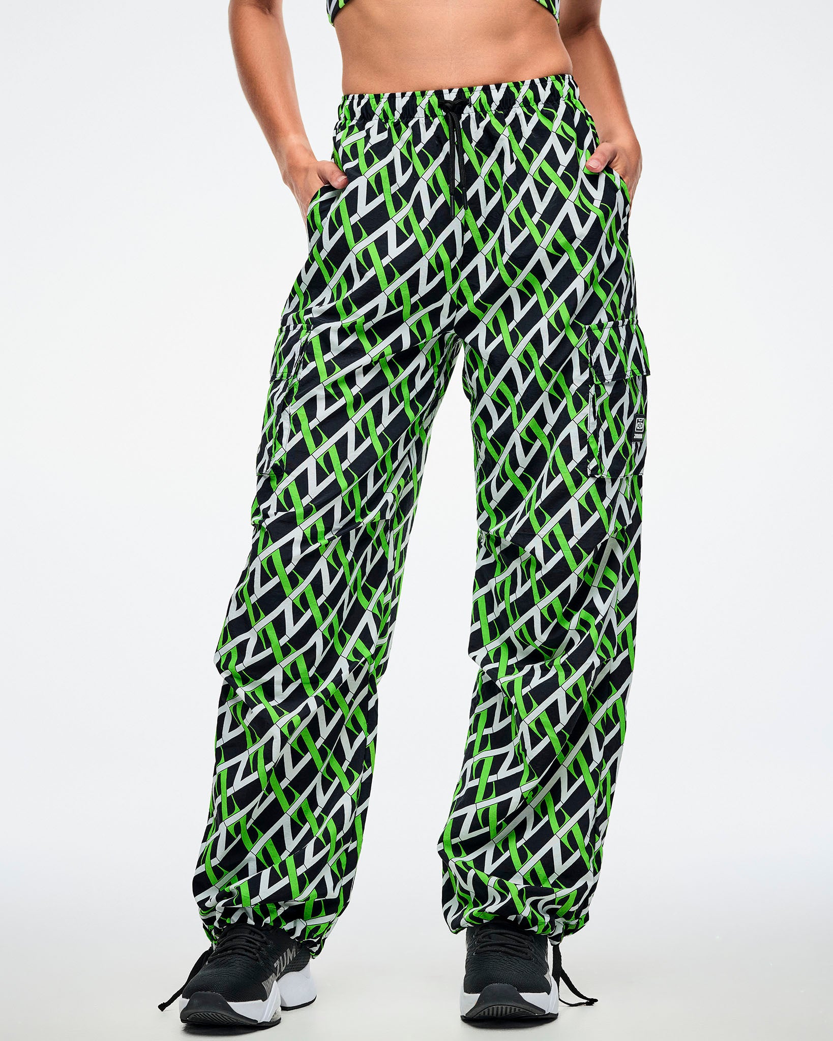 Front view of a female model wearing cargo pants with an allover black, white and neon green “Z” crisscross pattern, left hand in her pocket, paired with black-and-white shoes.