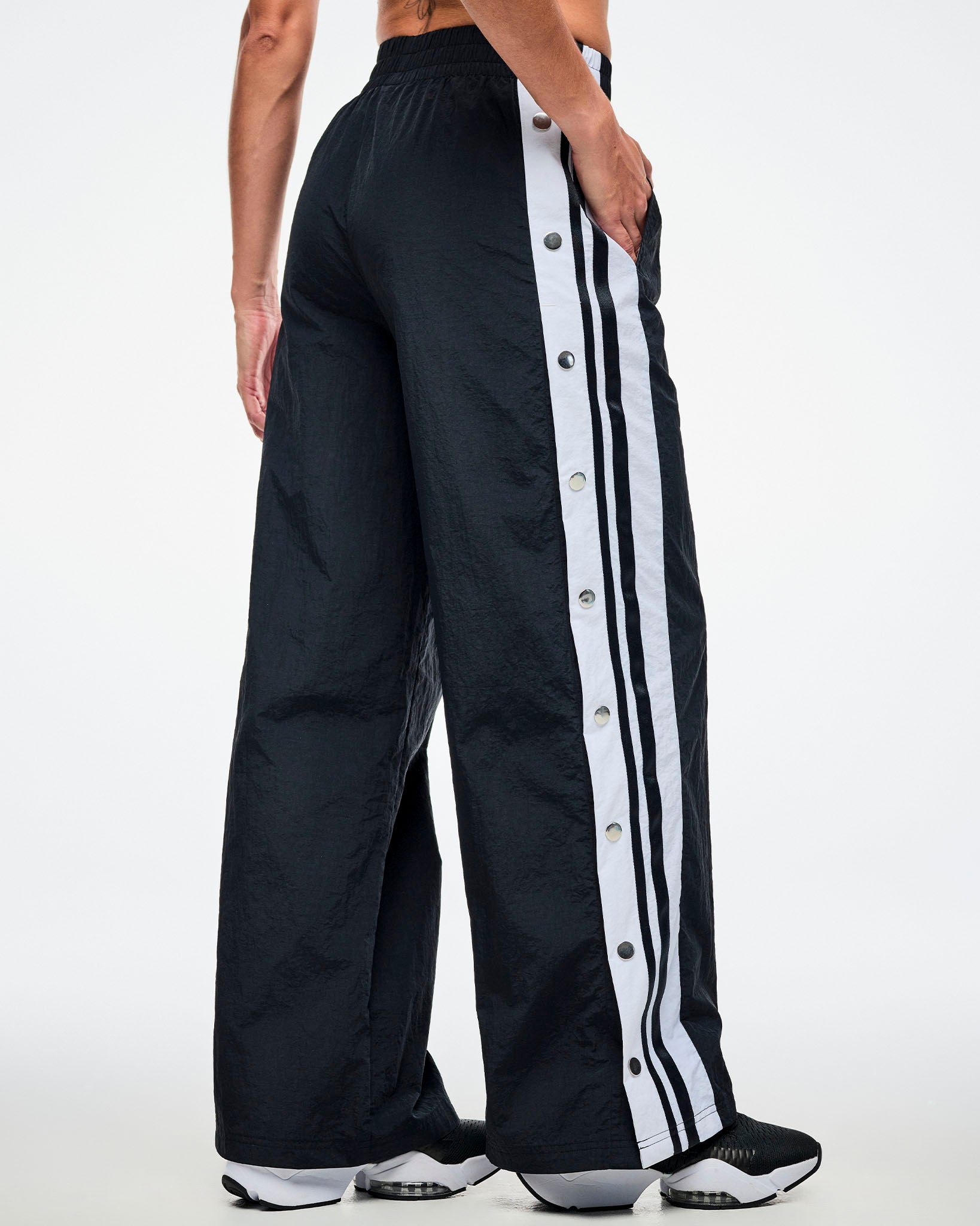 Back view of a female model wearing black pants with white stripes on the side with side snaps and paired with black and white shoes.