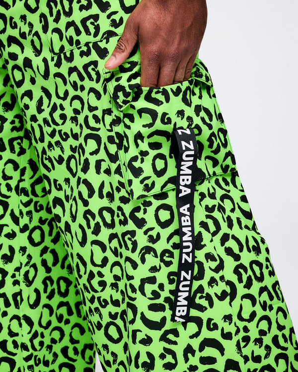Close-up of neon green leopard-print Zumba cargo pants, hand tucked into the cargo pocket. A vertical black tag along the pocket reads “Zumba