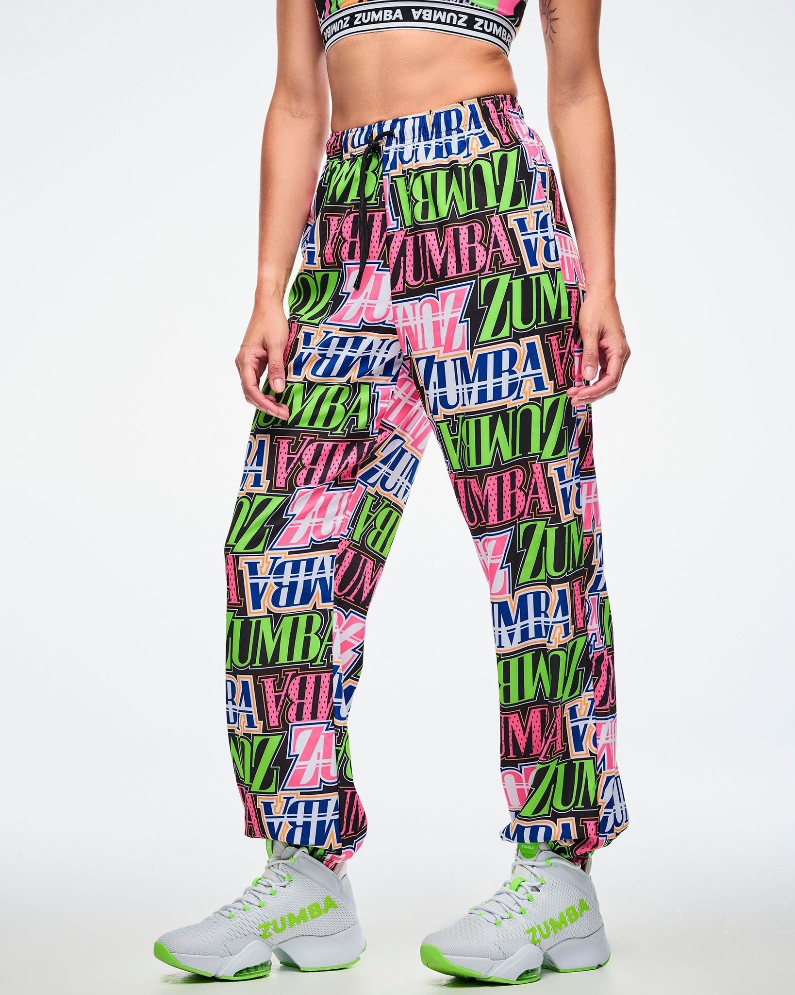 Female model wearing colorful Zumba pants paired with green and white shoes on a white background.