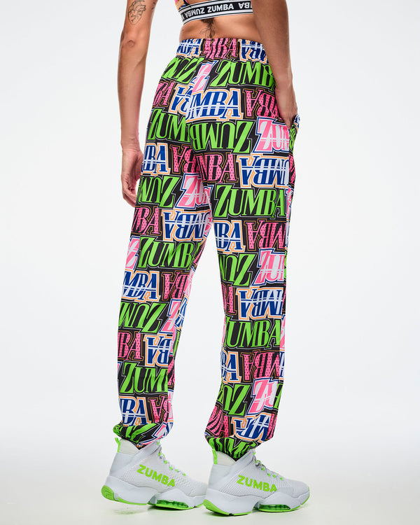 Back view of a female model wearing colorful Zumba pants paired with green and white shoes on a white background.