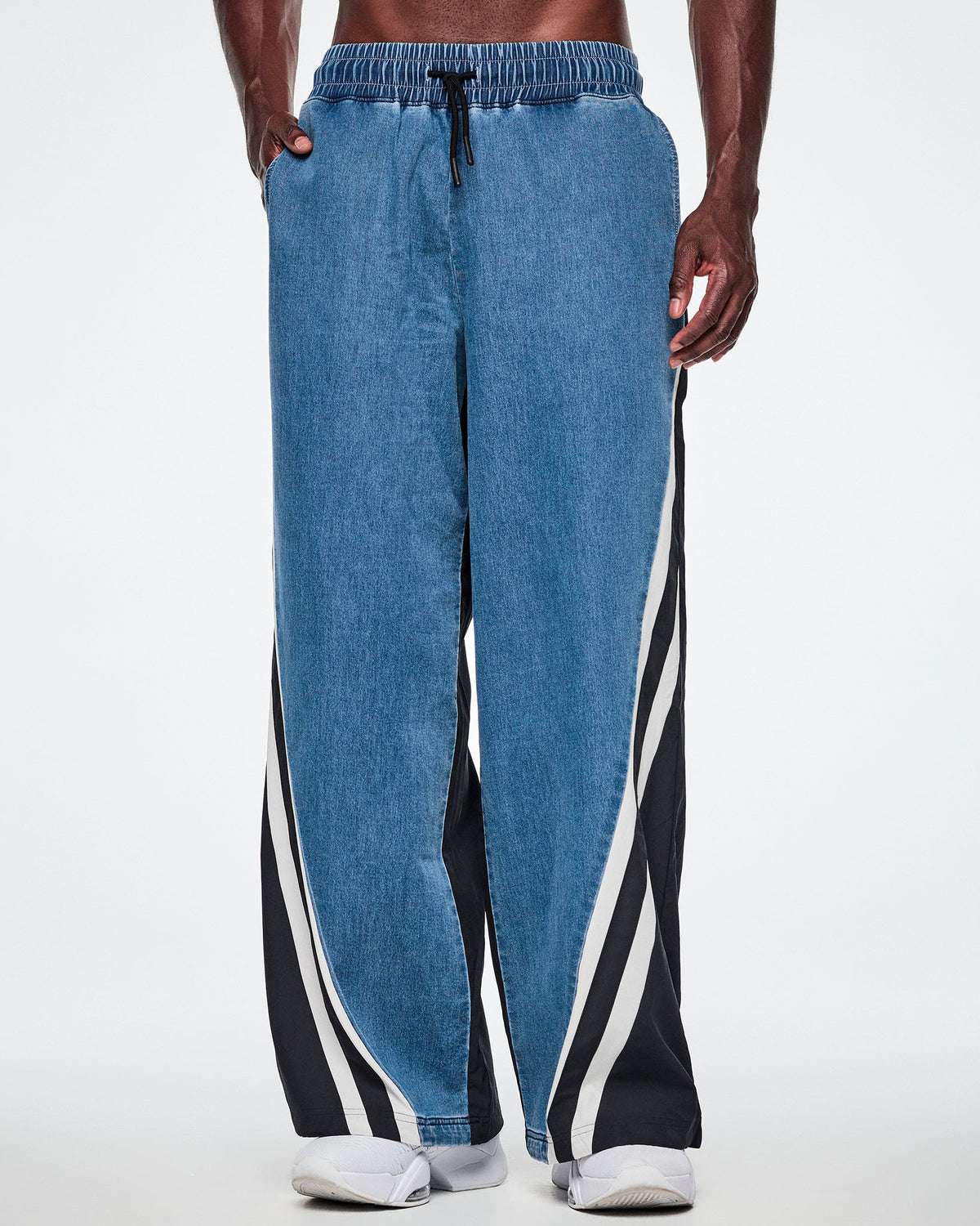 Front view of a male model wearing wide-leg denim combo pants with black-and-white striped side panels, paired with white sneakers.