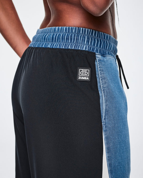 Close-up of the waistband on denim combo pants, showing the encased elastic waist with denim in front and black material in back, a drawcord, and a black tag featuring a cassette tape icon and 