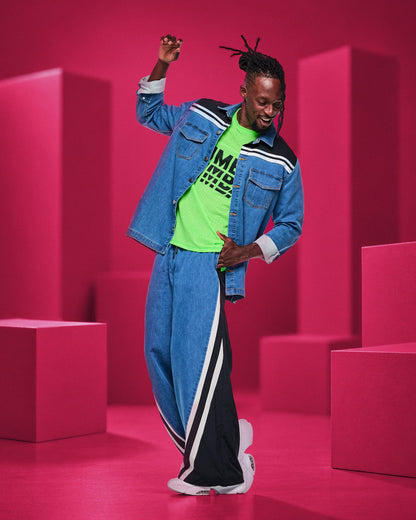 Male model dancing in a denim shirt jacket with a black shoulder yoke and white stripes, a neon green mesh top, and denim pants with a black panel and white stripes down the leg, set among pink blocks.