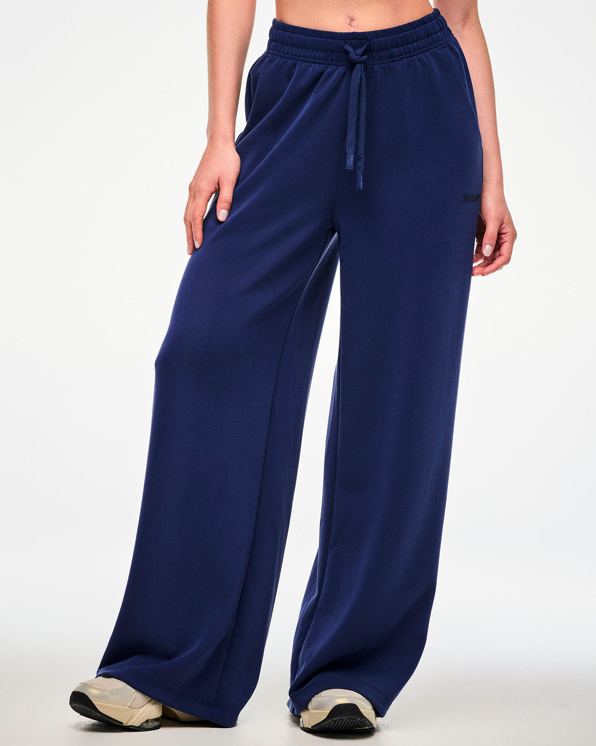 Female model in navy high-waisted straight-leg pants with a drawcord waist and tonal 'ZUMBA' text near the pocket on the left hip, paired with beige shoes.