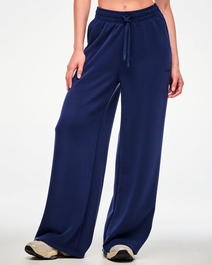 Female model in navy high-waisted straight-leg pants with a drawcord waist and tonal 'ZUMBA' text near the pocket on the left hip, paired with beige shoes.