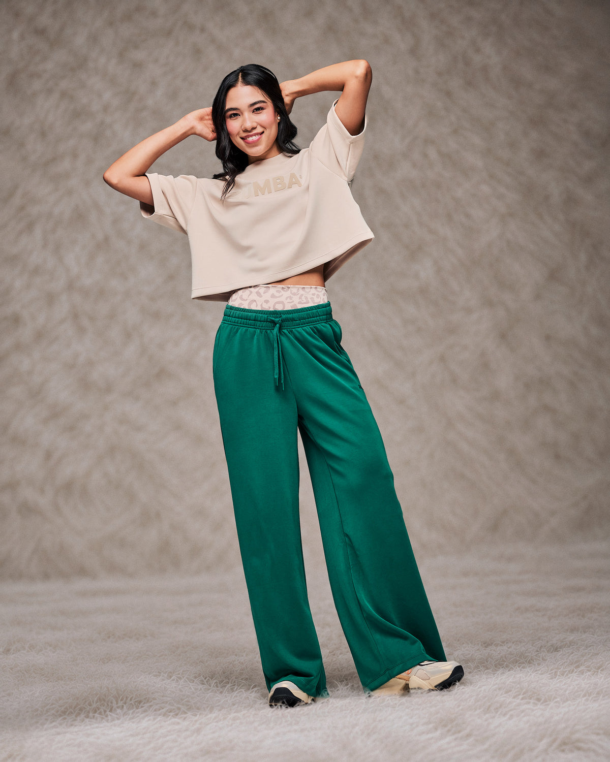Female model with both hands behind her head in the teal green high-waisted pants, styled with a beige drop shoulder crop top. Shot on a shaggy cream rug against a taupe faux fur backdrop.