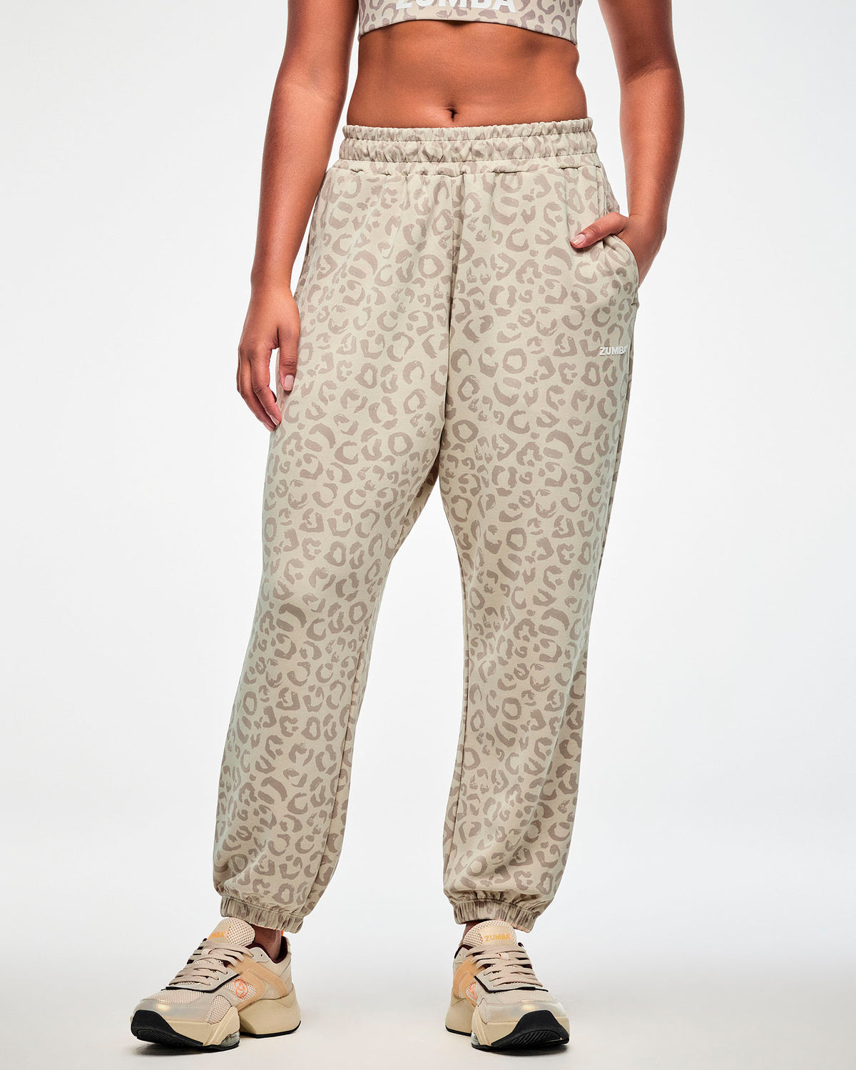 Female model in beige knit slouch cargo pants with an all-over light brown leopard print, white 'ZUMBA' text on the upper left leg, and elastic ankle cuffs, paired with a matching beige leopard print bra.