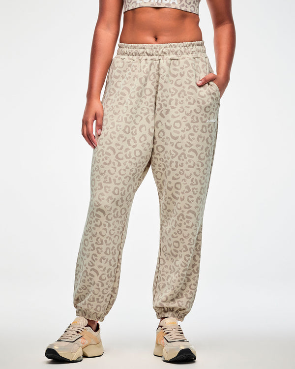 Female model in beige knit slouch cargo pants with an all-over light brown leopard print, white 'ZUMBA' text on the upper left leg, and elastic ankle cuffs, paired with a matching beige leopard print bra.