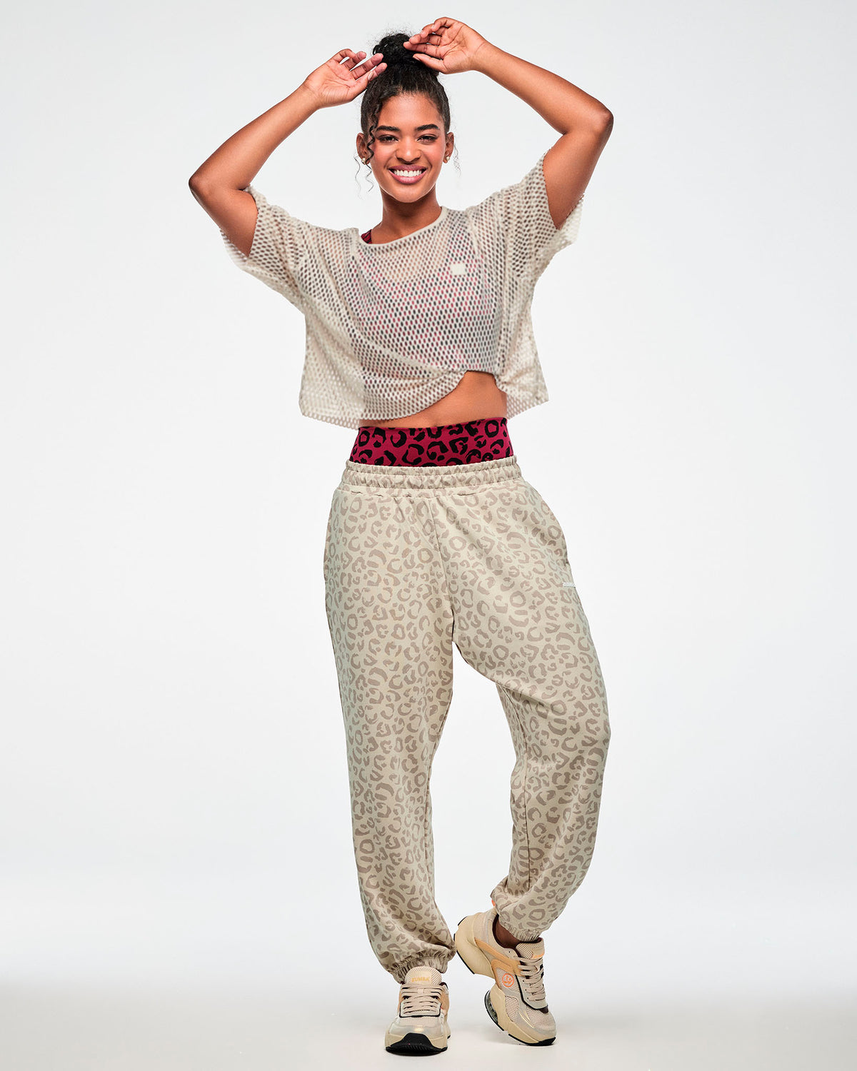 Full view of a female model with arms raised in the beige leopard print slouch cargo pants, styled with a beige mesh crop top over a dark red leopard print bra, dark red leopard print biker shorts layered underneath, and beige shoes.