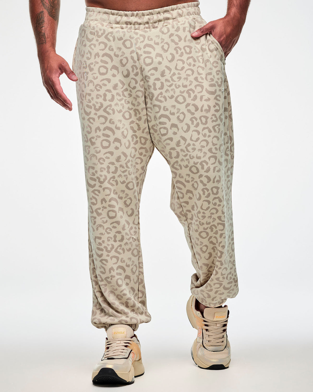 Male model in beige knit slouch cargo pants with an all-over light brown leopard print, elastic ankle cuffs, and one hand in the pocket, paired with beige shoes.