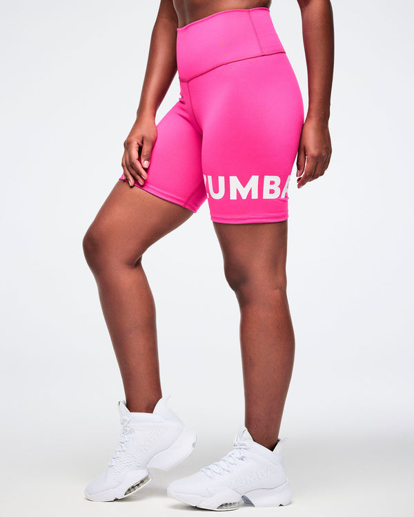 Side view of a woman wearing pink Zumba biker shorts with white Zumba branding on the left leg, styled with white shoes.