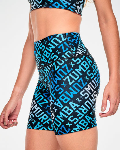 Female model in black high-waisted biker shorts with an all-over diagonal print of bold 'ZUMBA x THE SMURFS' varsity text in shades of blue and white on black, shown in side view to highlight the high-rise waistband. Paired with a matching print bra.