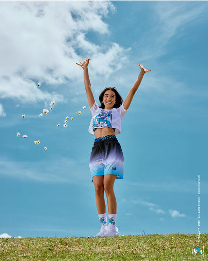 Female model with arms raised on a grassy hilltop, wearing the black-to-light-blue ombre mesh shorts with a white mesh Smurfs crop top and Smurf socks, as daisies fly across a bright blue sky.