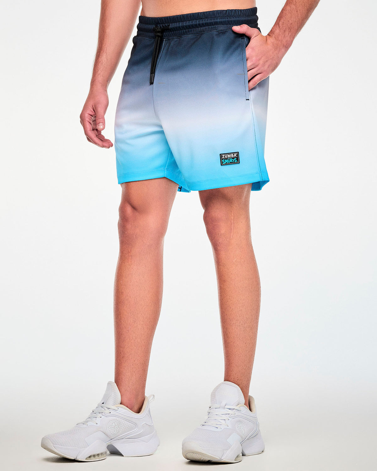 Angled view of a male model in navy-to-white-to-light-blue ombre mesh shorts, featuring an elastic waistband with a black drawcord and a black 'ZUMBA x THE SMURFS' patch near the left hem.
