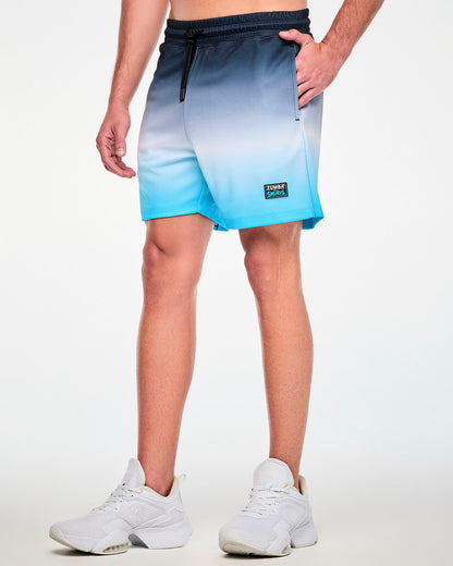 Angled view of a male model in navy-to-white-to-light-blue ombre mesh shorts, featuring an elastic waistband with a black drawcord and a black 'ZUMBA x THE SMURFS' patch near the left hem.