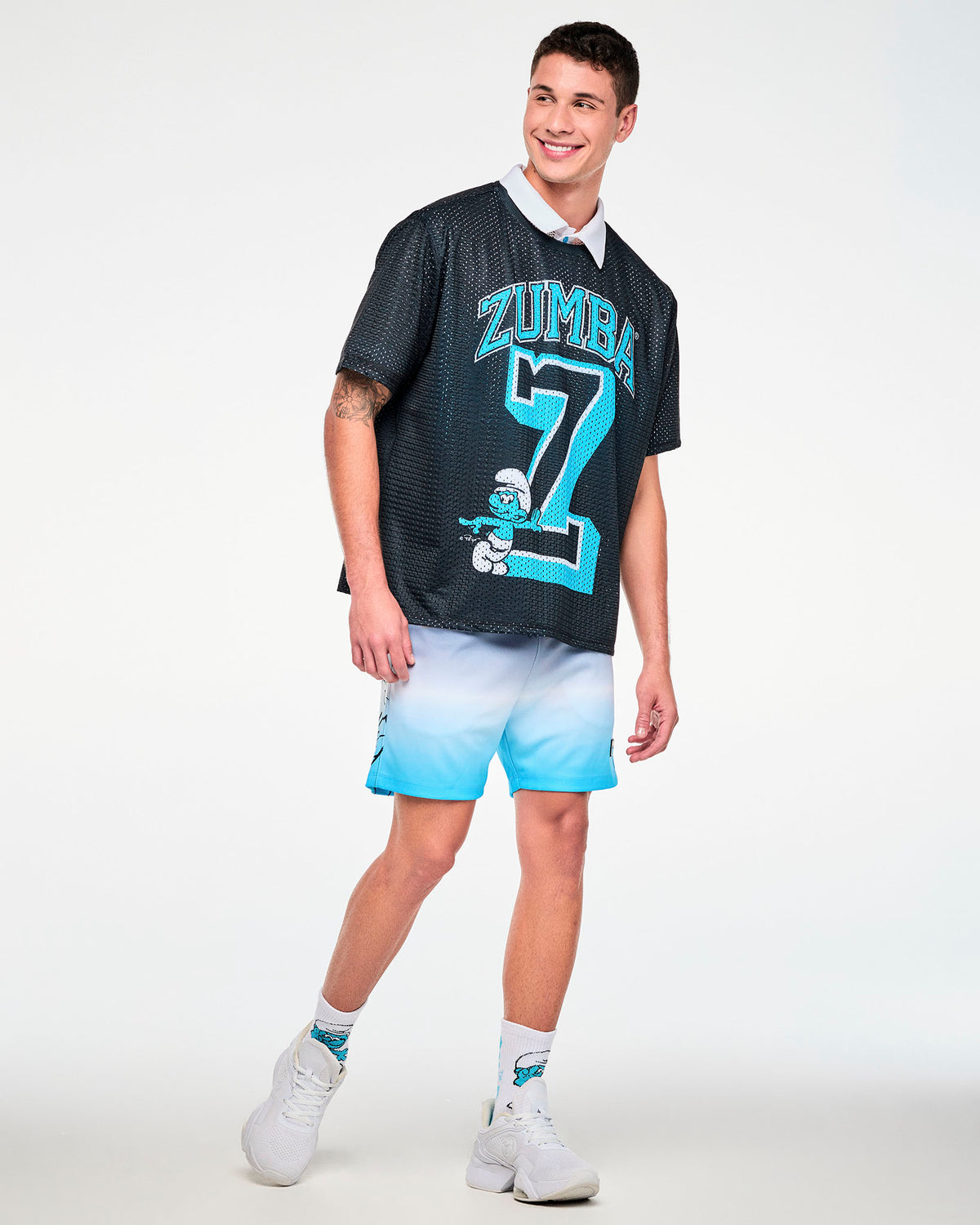 Male model in the navy-to-light-blue ombre mesh shorts, with the outline Smurf graphic near the hem of the right leg, paired with a black mesh boxy tee with light blue 'ZUMBA' text and a varsity 'Z' over a white collar, Smurf socks, and white shoes.