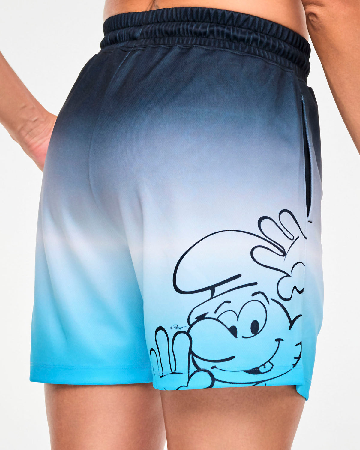 Close-up of the back of the navy-to-light-blue ombre mesh shorts, showing the navy line drawing of a Smurf peeking up with a raised hand, and the pocket opening at the side.