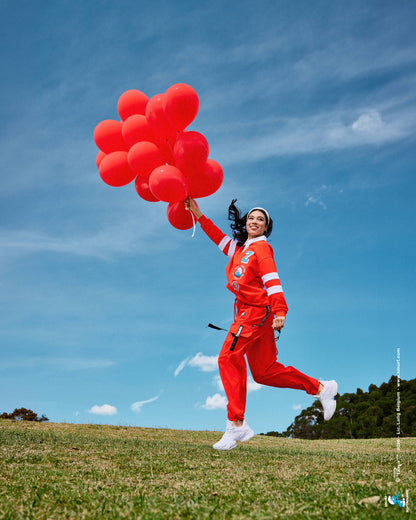 Female model running across a grassy hill holding a bunch of red balloons, wearing the red cargo pants with black 'ZUMBA' straps and the red rugby 'ZUMBA' and 'THE SMURFS' polo under a blue sky.