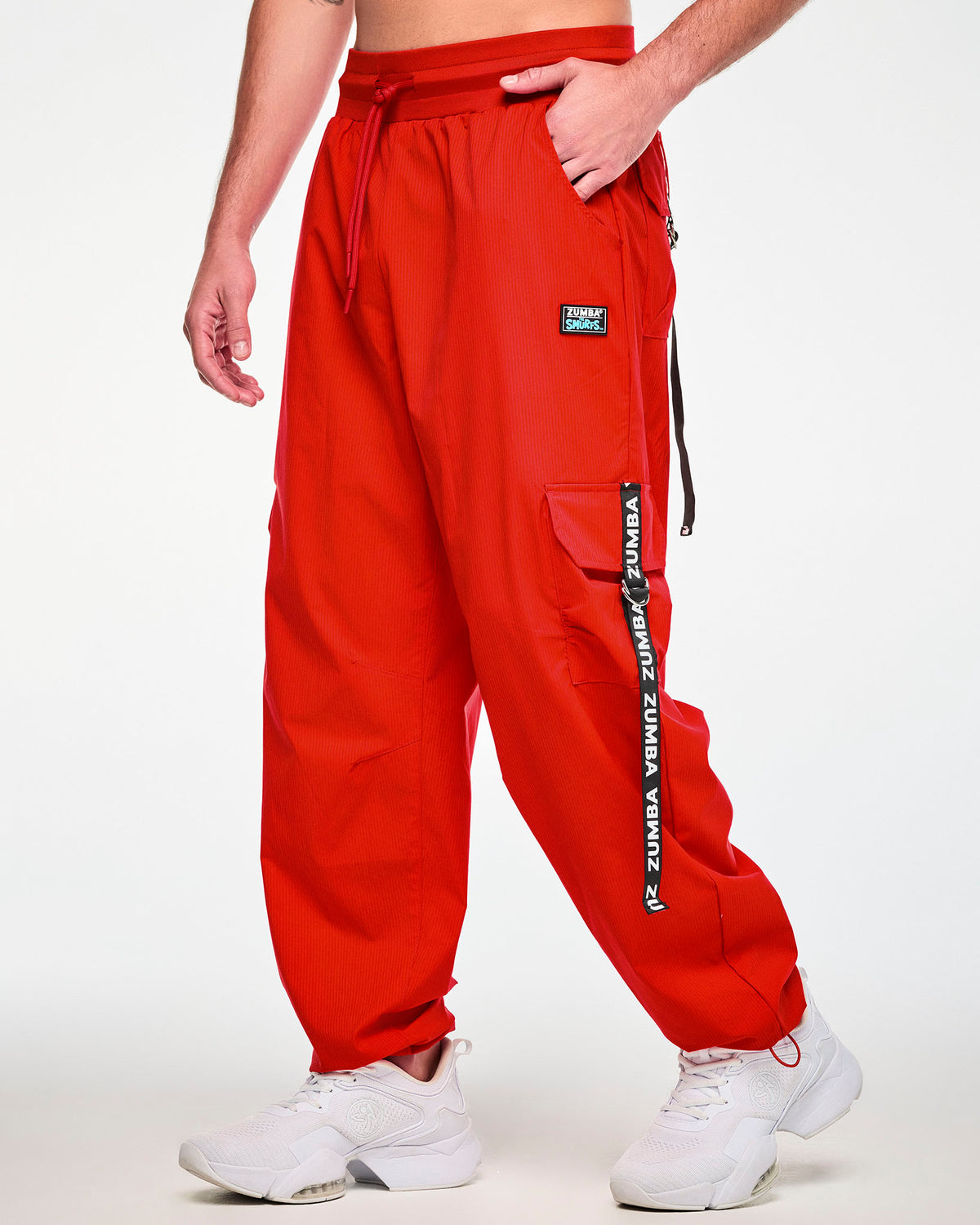 Male model with one hand in the pocket of red woven cargo pants with a fine tonal stripe texture, shown in side view with a black 'ZUMBA' and 'THE SMURFS' patch on the left hip and a black 'ZUMBA' webbing strap looped through a silver D-ring on the cargo pocket.