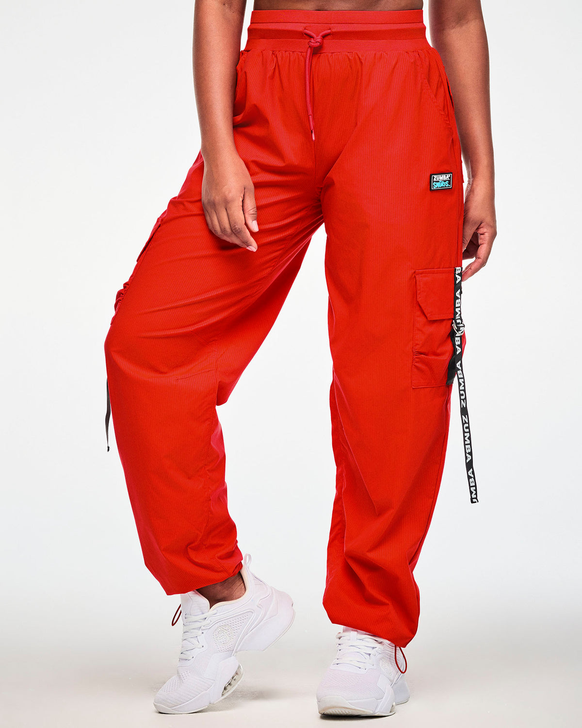 Female model in the red cargo pants, showing the wide waistband with a red drawcord, the black 'ZUMBA' X 'THE SMURFS' patch on the left hip, and a black 'ZUMBA' webbing strap on the cargo pocket. Red bungee cords cinch the cuffs over white shoes.