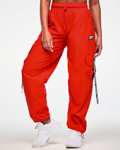 Female model in the red cargo pants, showing the wide waistband with a red drawcord, the black 'ZUMBA' X 'THE SMURFS' patch on the left hip, and a black 'ZUMBA' webbing strap on the cargo pocket. Red bungee cords cinch the cuffs over white shoes.