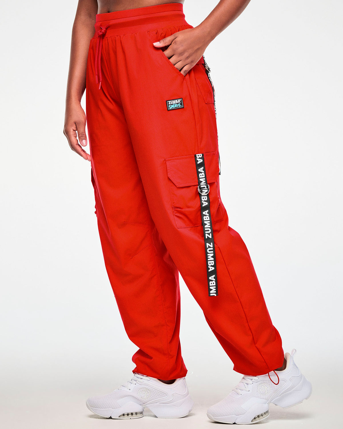 Female model with one hand in the pocket of red woven cargo pants with a fine tonal stripe texture and a loose, relaxed leg, shown in side view with a black 'ZUMBA' webbing strap hanging from the cargo pocket past the knee.