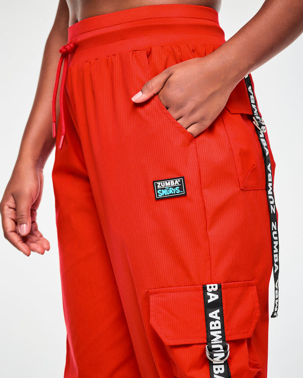 Close-up of the red cargo pants at the hip, showing the drawcord, the 'ZUMBA' X 'THE SMURFS' patch, and 'ZUMBA' webbing straps with silver D-rings on the cargo pockets.
