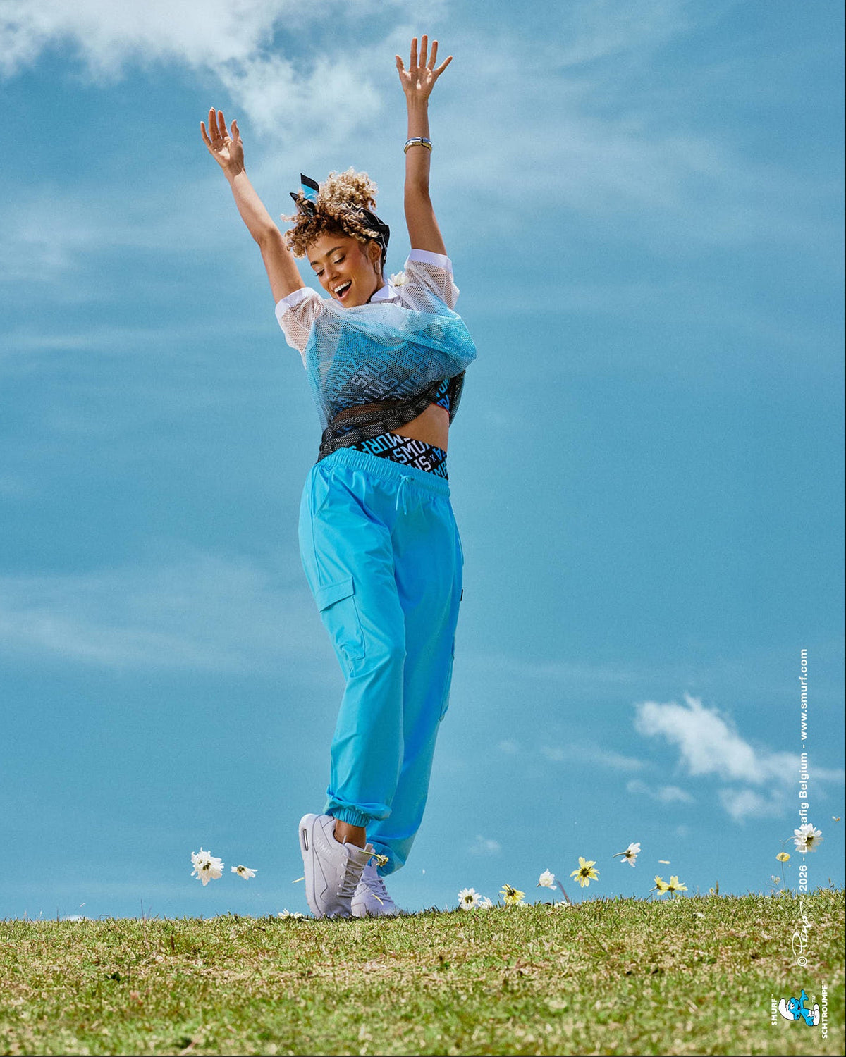 Female model dancing on a grassy hilltop with arms raised, wearing aqua blue baggy cargo pants with a loose, relaxed fit, paired with a mesh polo and logo print bra, with daisies scattered in the grass under a cloudy blue sky.