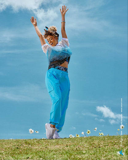 Female model dancing on a grassy hilltop with arms raised, wearing aqua blue baggy cargo pants with a loose, relaxed fit, paired with a mesh polo and logo print bra, with daisies scattered in the grass under a cloudy blue sky.