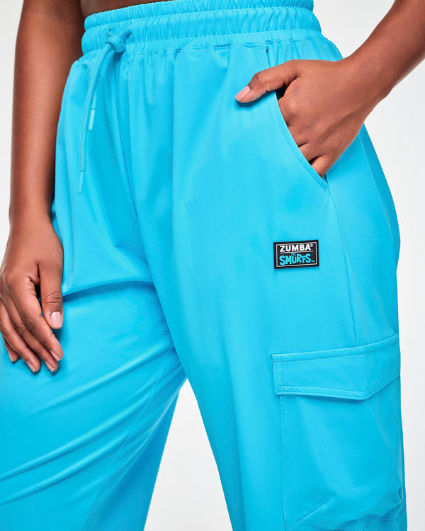 Close-up of the aqua blue cargo pants, showing the gathered elastic waistband, drawcord, slanted pocket, 'ZUMBA' X 'THE SMURFS' patch, and the flap cargo pocket.